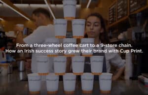 Shown is a pyramid of Coffee Angel custom branded paper takeaway coffee cups in customer environment. Image is for a Coffee Angel customer testimonial discussing custom printed cups from Cup Print, and has text on it saying 'From a 3-wheel coffee cart to a 9-store chain. How an Irish success story grew their brand with Cup Print'